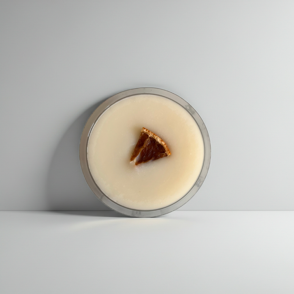 A round glass jar filled with a creamy off-white product, featuring a small triangular piece of dark brown and amber citrus peel on top, set against a minimalist light gray background.