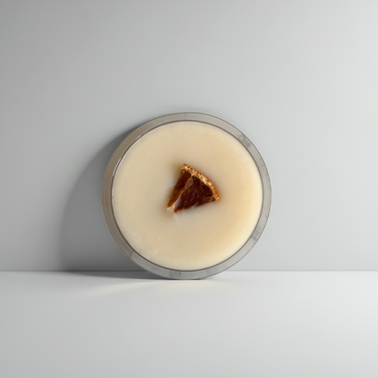 A round glass jar filled with a creamy off-white product, featuring a small triangular piece of dark brown and amber citrus peel on top, set against a minimalist light gray background.
