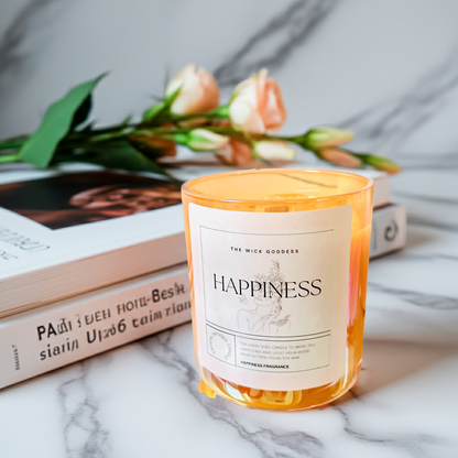 Candle labeled 'Happiness' on a marble surface with books and flowers in the background